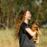 Young woman joyfully holding her Norwich terrier dog outdoors in a sunlit park.